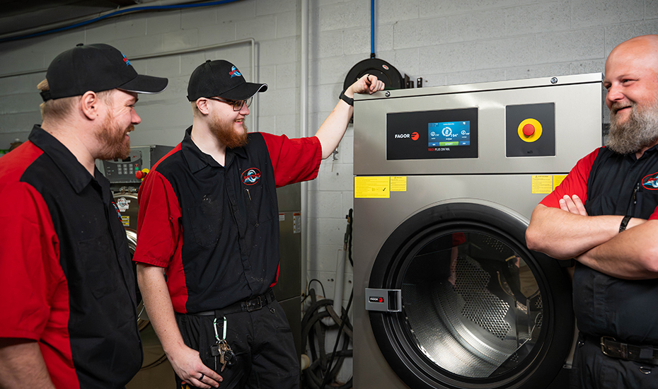 Three workers in red-and-black uniforms stand beside a large industrial front-loading washer, chatting by the control panel.