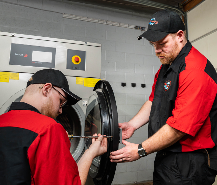 Two technicians in red and black uniforms work on an open industrial machine, tightening a cylindrical component with a torque wrench.